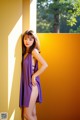 A woman in a purple dress leaning against a yellow wall.